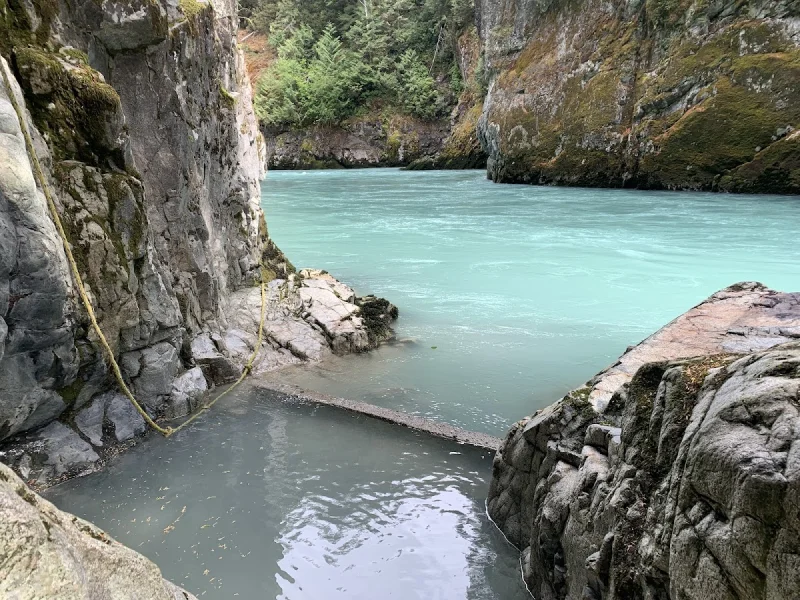 Rugged rock hot spring pool beside a turquoise river in forested canyon