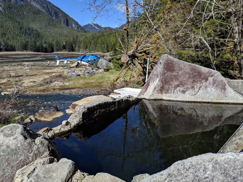 Remote hot springs pool with helicopter landing area and forested mountain backdrop